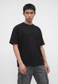 Junger Mann trägt ein schlichtes schwarzes oversized T-Shirt und graue Cargohosen und steht vor einem weißen Hintergrund.