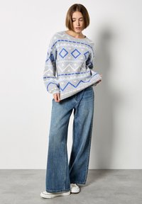 Maglione grigio lavorato a maglia con motivi a diamante e zigzag blu, abbinato a jeans in denim blu a gamba larga e sneaker bianche. Collo giro semplice.