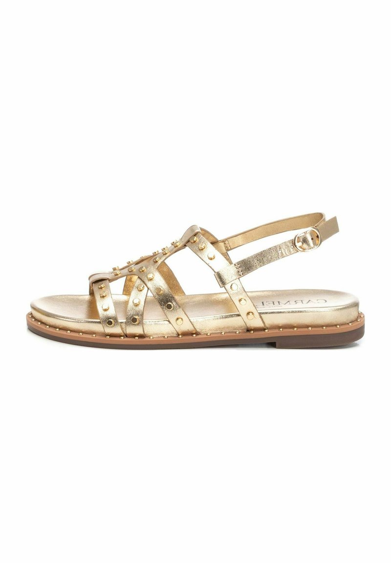 Sandalias metálicas doradas con correas con tachuelas, suela plana, correa de tobillo ajustable y textura suave. Herrajes decorativos a lo largo del borde.