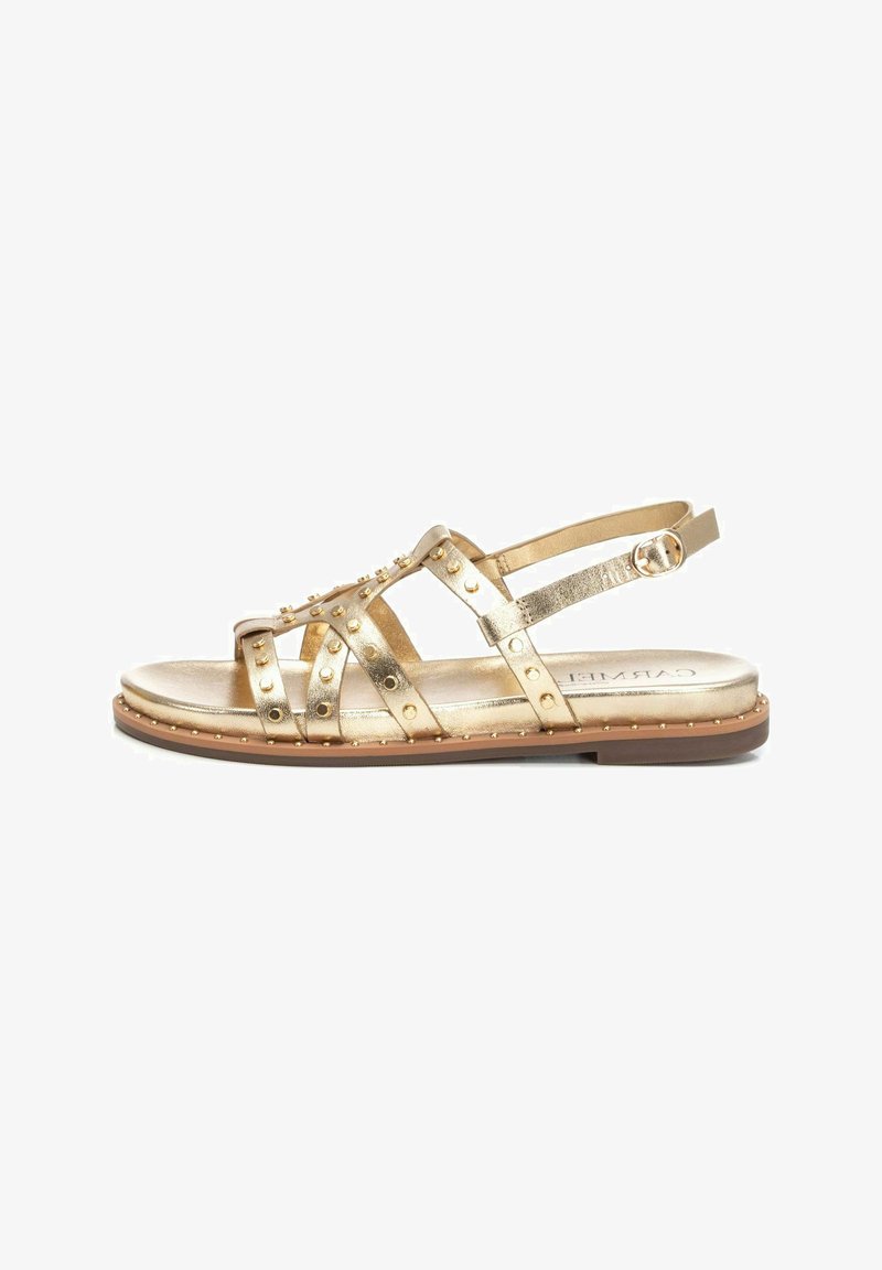 Sandalias metálicas doradas con correas con tachuelas, suela plana, correa de tobillo ajustable y textura suave. Herrajes decorativos a lo largo del borde.