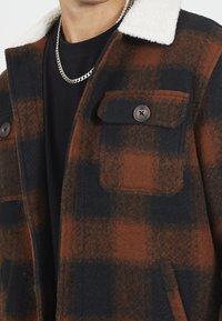 Brun och svart rutigt jacka med en vit fuskpäls krage, som har två framfickor och stora knappar, tillverkad av en texturerad ullblandning.