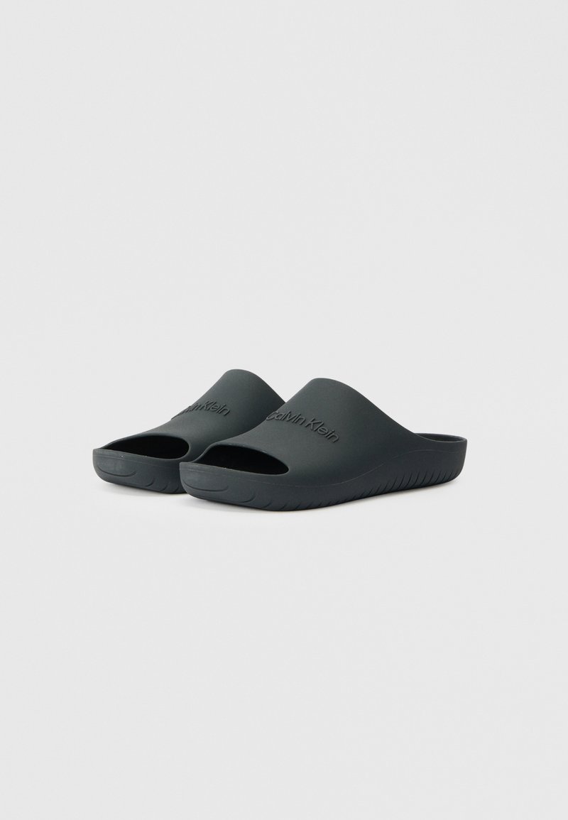 Schwarze Gummischuhe mit offenem Zehendesign, ausgestattet mit einer strukturierten Sohle und eingeprägtem Logo auf dem oberen Riemen. Glatte Oberfläche und minimalistischer Stil.