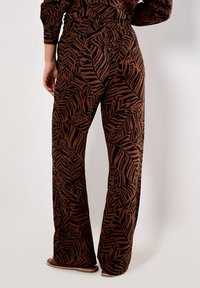 Brown wide-leg pants featuring a black leaf pattern, elastic waistband, and a smooth fabric texture. Worn with flat sandals.