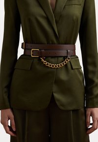 Brun läderbälte med ett guld kedjeaccent, bärs över en olivgrön blazer, vilket framhäver en strukturerad design och slät textur.