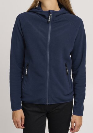 Frau trägt eine marineblaue Fleecejacke mit Reißverschluss und Kapuze sowie schwarze Hose, steht mit entspannt an den Seiten hängenden Armen.