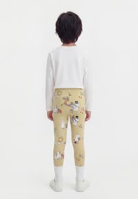 Παιδί που κοιτάζει προς τα πίσω, φοράει μπεζ κολάν με φανταστικούς καρτούν χαρακτήρες και λευκό μπλουζάκι με μακριά μανίκια, λευκές κάλτσες και λευκά παπούτσια.