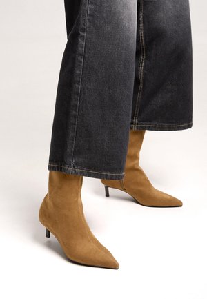 Bottines en daim couleur camel à bout pointu avec talons kitten, portées sous un jean noir large et raccourci, sur un fond clair.