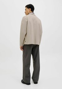 Beige jas met een relaxte pasvorm, zachte textuur en zijsplitten, gecombineerd met loszittende donkergrijze broek en zwarte schoenen.
