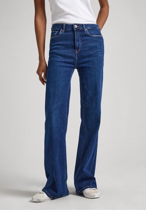 Personne portant un jean bleu large, un haut blanc sans manches et des baskets blanches, debout devant un fond uni clair.
