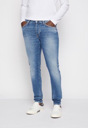Personne portant un jean en denim bleu clair et des baskets blanches, les mains dans les poches, debout devant un fond blanc uni.