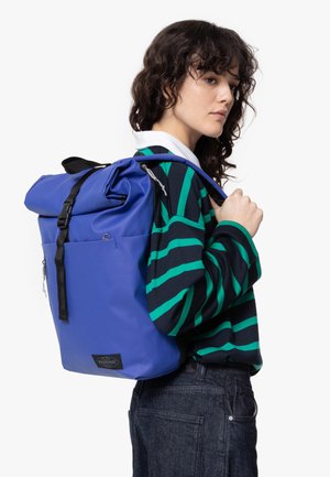 Mochila azul con cierre enrollable, hecha de material duradero, con correas y cremalleras negras, un bolsillo frontal y etiqueta de marca.
