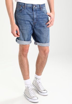 Homme portant un short en denim bleu retroussé, des baskets blanches montantes, des chaussettes blanches et une chemise blanche, debout devant un fond uni.