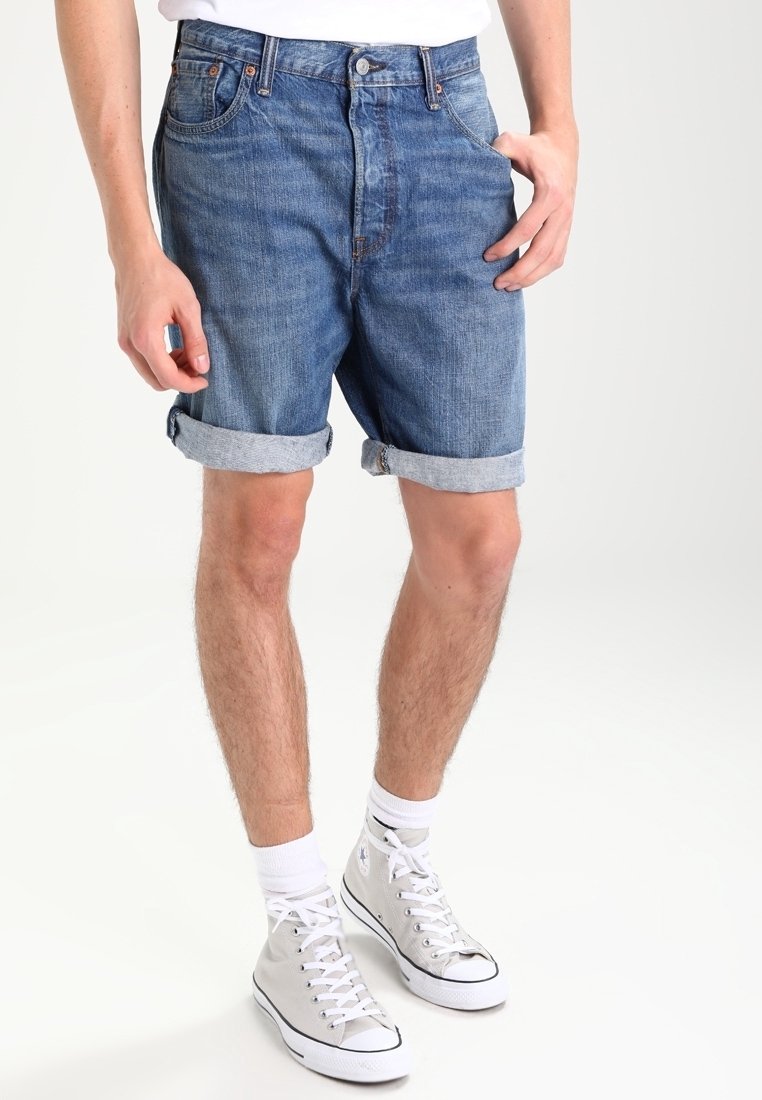 Homme portant un short en denim bleu retroussé, des baskets blanches montantes, des chaussettes blanches et une chemise blanche, debout devant un fond uni.