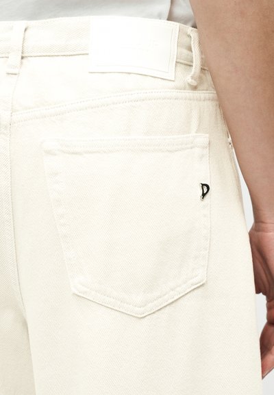 Gros plan sur une personne portant un jean en denim écru avec une poche arrière ornée d'un petit logo métallique noir.