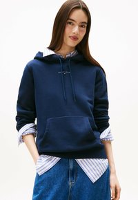 Felpa navy con tasca frontale, cordoncino regolabile e logo. Indossata sopra una camicia a righe blu chiaro; abbinata a jeans.