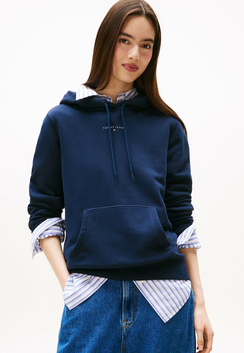 Felpa navy con tasca frontale, cordoncino regolabile e logo. Indossata sopra una camicia a righe blu chiaro; abbinata a jeans.