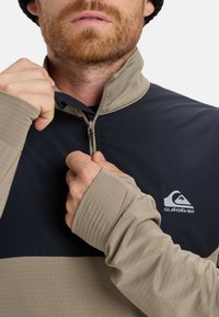 Quarter-zip trøje i beige og navy, med en glat tekstur, teksturerede ærmer og et logo på brystet. Høj krave detalje synlig.