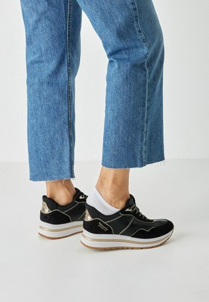 Schwarze Sneaker aus Wildleder und Lack mit weißer Sohle, metallischen Akzenten, Schnürsenkeln und leicht erhöhtem Absatz. Kombiniert mit hochgekrempelten blauen Jeans.