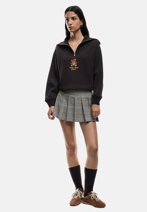 Bruine zip-up hoodie met een teddybeer graphic, gecombineerd met een geplooide geruite rok. Model draagt bruine schoenen en kniekousen.