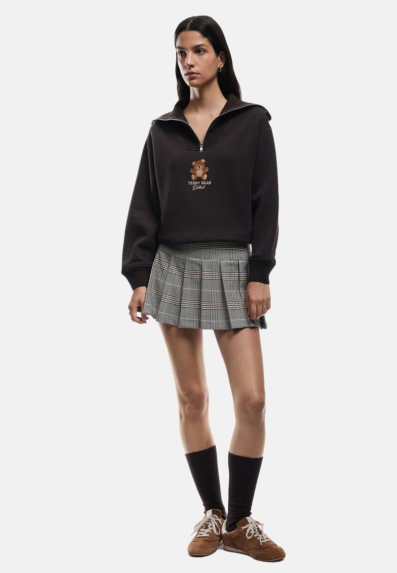 Brązowy hoodie na zamek z grafiką misia, w zestawieniu z plisowaną spódnicą w kratkę. Modelka ma na sobie brązowe buty i skarpetki do kolan.