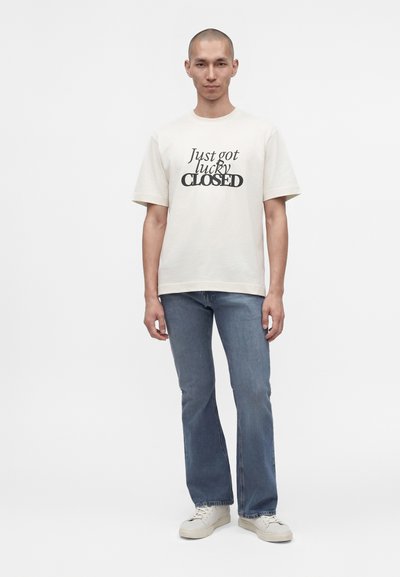 Homme portant un t-shirt blanc avec le texte ""Just got lucky CLOSED"", un jean bleu et des baskets blanches, debout devant un fond uni.