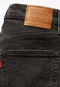 Svarte denimjeans med en teksturert overflate, med et brunt lærmerke med preget branding og sømdetaljer langs lommene.