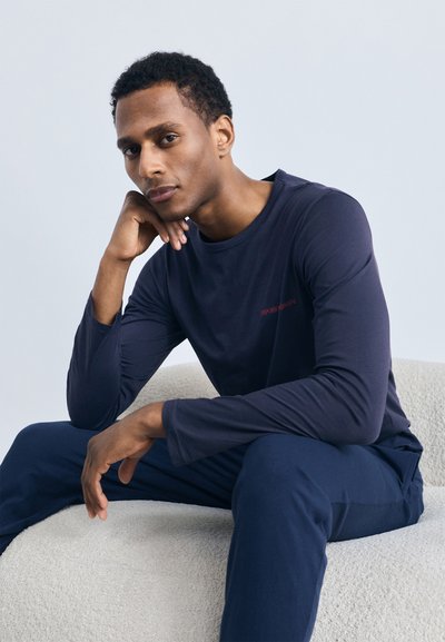 Navy long-sleeve shirt with round neckline, featuring subtle red logo detail. Model seated on a textured, light-colored surface.