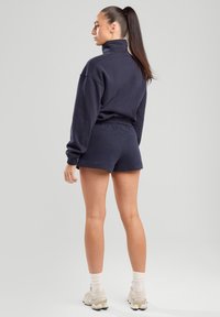 Tummansininen collegepaita korkealla kauluksella, yhdistettynä yhteensopiviin shortseihin. Pehmeä kangas, rento muotoilu, näkyvät saumat ja käytetään kevyiden tennareiden kanssa.