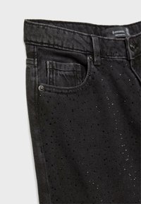 Črne denim hlače z teksturiranim zaključkom, ki imajo majhne, sijoče črne dodatke. Vključujejo sprednji žep in kovinske gumbe.