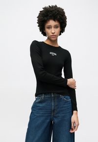 Top preto de manga comprida com textura canelada e gola redonda, com o logo branco "Guess" no peito. Combinado com jeans azuis largos.