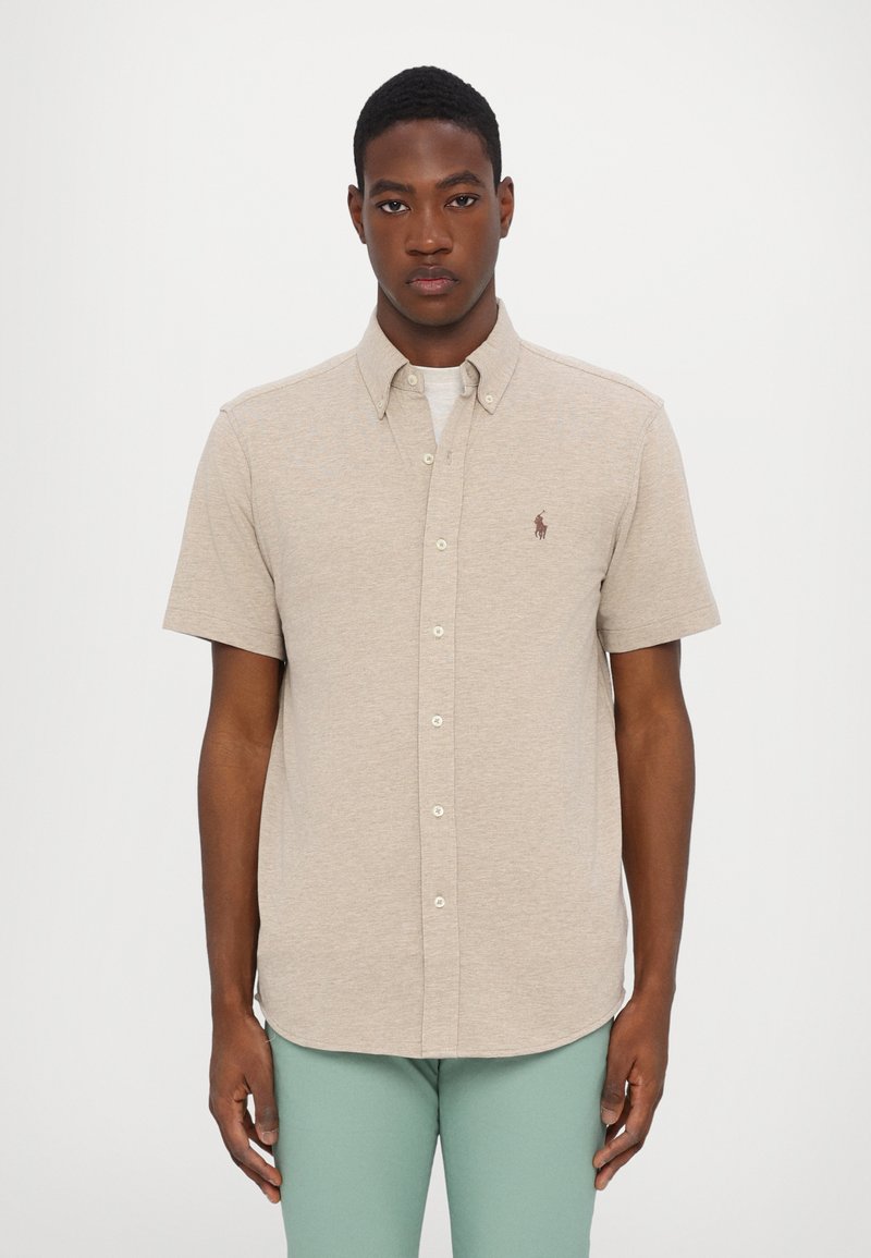 Camisa beige de manga corta con cuello abotonado, que presenta una tela texturizada y un pequeño logo en el pecho.