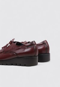 Zapatos de cuero burdeos con puntera redondeada, suela negra gruesa y adornos de borlas en el frente. Presentan una pequeña etiqueta de la marca en el costado.