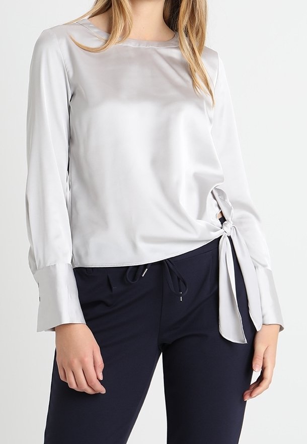 Blusa in satin argento con maniche lunghe, scollatura rotonda, dettaglio nodo laterale e texture liscia. Abbinata a pantaloni blu scuro.