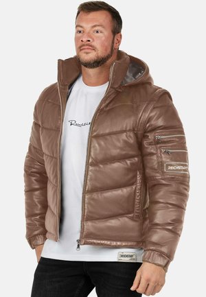 Man draagt een bruine gewatteerde leren jas met capuchon, rits aan de voorkant en meerdere ritsen aan de linker mouw, over een wit shirt met zwarte tekst, staand.