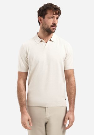 Mann trägt ein beiges kurzärmeliges Poloshirt und helle Hose, schaut mit neutralem Ausdruck leicht nach rechts.