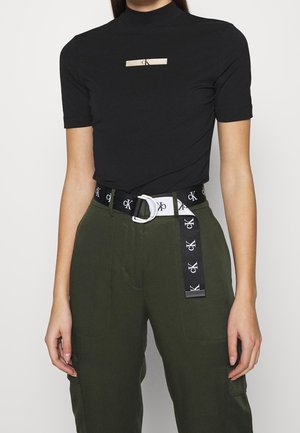 Kobieta w czarnym krótkim topie z małym logo oraz oliwkowych spodniach z wysokim stanem z czarno-białym paskiem z logo Calvin Klein.