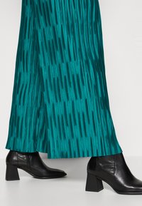 Falda larga plisada en color azul verdoso con textura vertical, combinada con botines de tacón negro, con punta cuadrada y acabado en cuero suave.