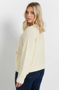 Gräddfärgad stickad tröja med en texturerad design, ribbade ärmar och en framficka. Bärs med mörkblå jeans.