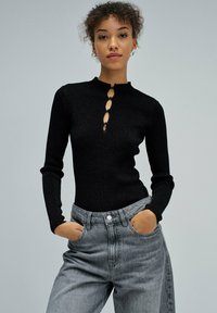 Maglia nera a maniche lunghe con dettaglio a occhiello e bottone sul collo. Abbinata a jeans in denim grigi chiari a vita alta.