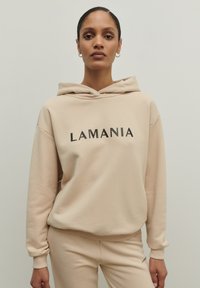 Béžová mikina s kapucňou, na ktorej je na prednej strane čiernym písmom vytlačené "LAMANIA"; rebrované manžety a pás; hladká textúra; skombinovaná s rovnakými béžovými nohavicami.