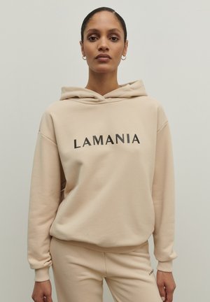 Béžová mikina s kapucňou, na ktorej je na prednej strane čiernym písmom vytlačené "LAMANIA"; rebrované manžety a pás; hladká textúra; skombinovaná s rovnakými béžovými nohavicami.