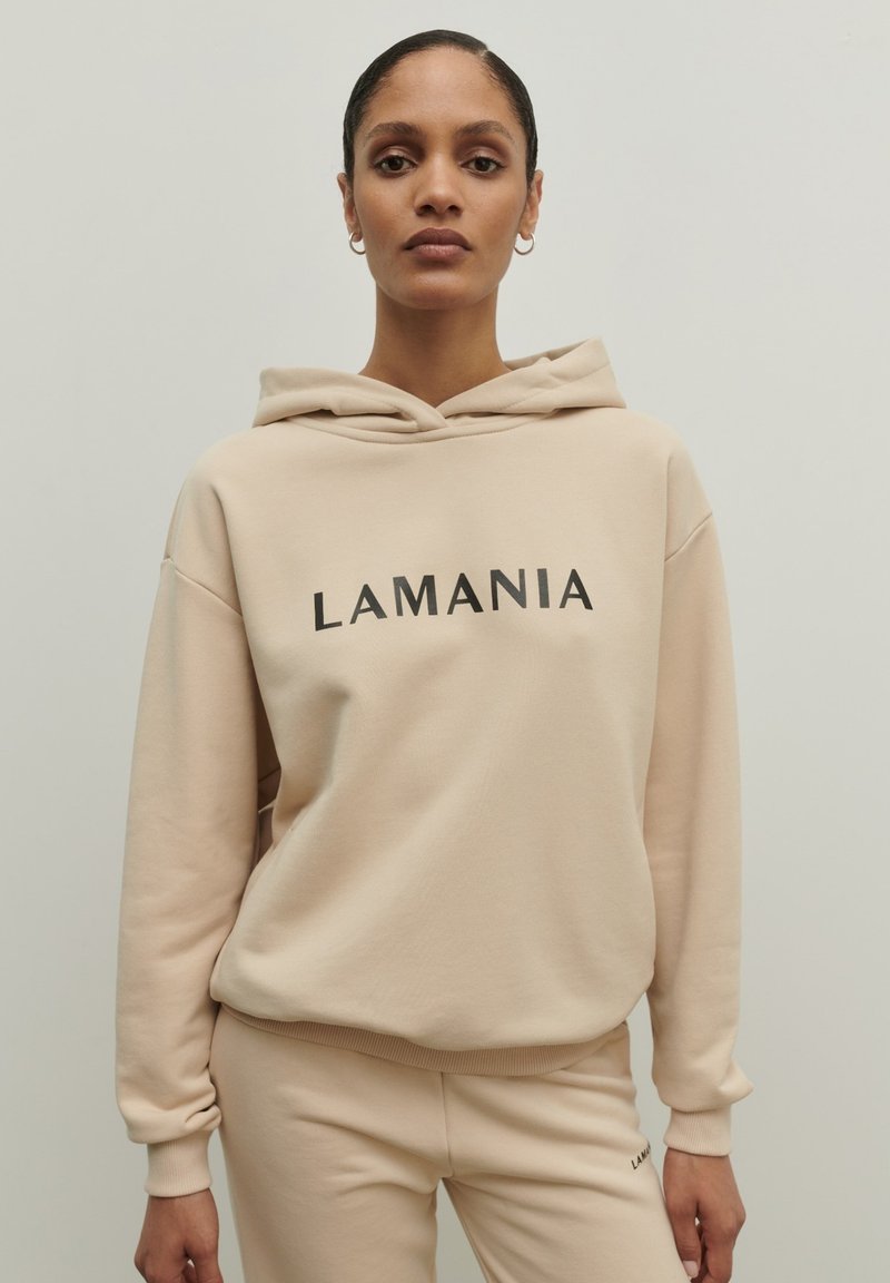 Béžová mikina s kapucňou, na ktorej je na prednej strane čiernym písmom vytlačené "LAMANIA"; rebrované manžety a pás; hladká textúra; skombinovaná s rovnakými béžovými nohavicami.