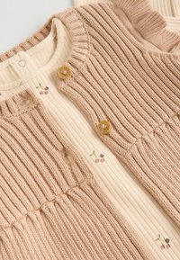 Cárdigan de punto acanalado en rosa claro, con botones de madera, combinado con una blusa crema que presenta un sutil bordado de cerezas y un patrón texturizado.