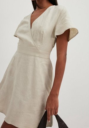 Vrouw draagt een beige linnen wikkeljurk met korte mouwen, houdt een donker gestructureerde handtas vast tegen een effen achtergrond.