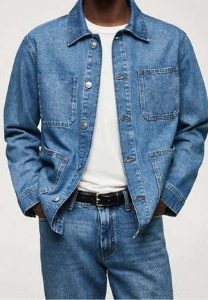 Homme portant une veste en jean bleue et un jean sur un t-shirt blanc, les mains dans les poches de la veste.