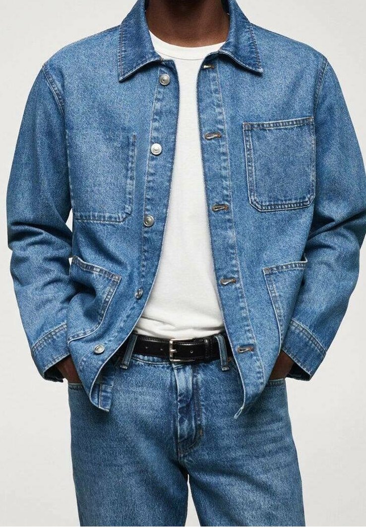 Homme portant une veste en jean bleue et un jean sur un t-shirt blanc, les mains dans les poches de la veste.