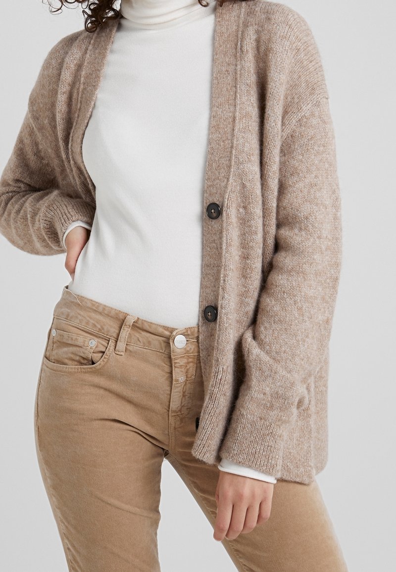 Beige gebreide cardigan met grote knopen, met een losse pasvorm en een textuurstof. Draagt over een witte coltrui en gecombineerd met beige broek.