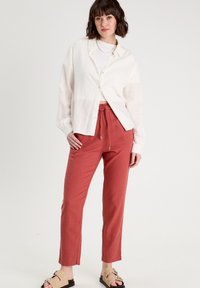 Chemise blanche à boutons et à manches longues, associée à un pantalon en cordon de couleur rouille. Sandales beiges à lanières avec des accents métalliques.
