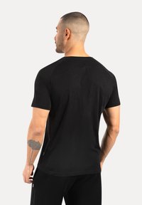 Man met kort haar, gekleed in een eenvoudig zwart T-shirt met korte mouwen en zwarte broek, toont een bloemen tattoo op zijn linker onderarm.