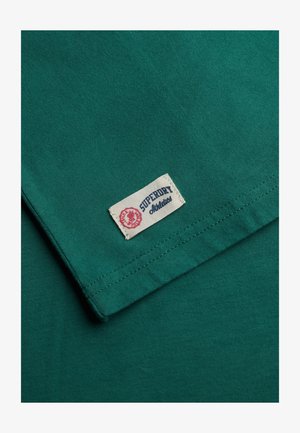Groen katoenen T-shirt met een Superdry-label. De stof is glad met een lichte glans en toont nette stiksels langs de zoom.